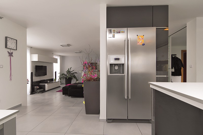 Americká chladnička je součástí polopříčky, která opticky odděluje kuchyň od vstupní a komunikační zóny přízemí. Propojenost jednotlivých zón interiéru je podpořena velkoformátovou šedou dlažbou.