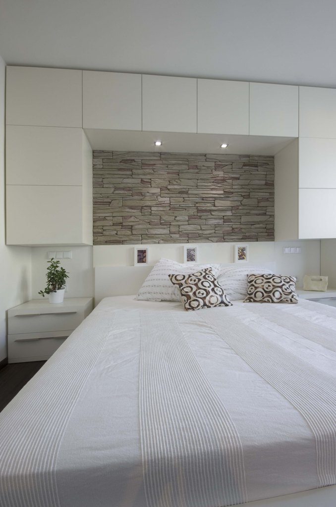 Světlá ložnice s barevností tón v tónu. Kamenný obklad nad postelí je možné večer nasvítit a vytvořit tak intimní atmosféru.