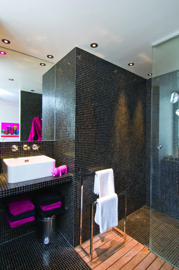 Koupelna působí exoticky díky stěnám a podlaze, obloženým lesklou černou mozaikou. Pocit tepla dodávají dřevěný rošt před sprchou a barevné textilie.