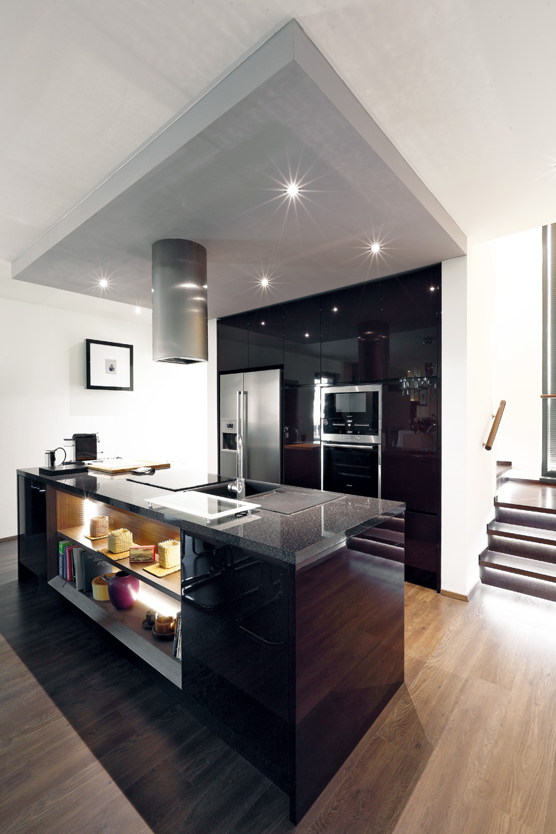 Kuchyň navzdory propojení s obývací částí působí kompaktně díky sníženému sádrokartonovému stropu. Vysoké skříně využívají niku vytvořenou schodištěm.