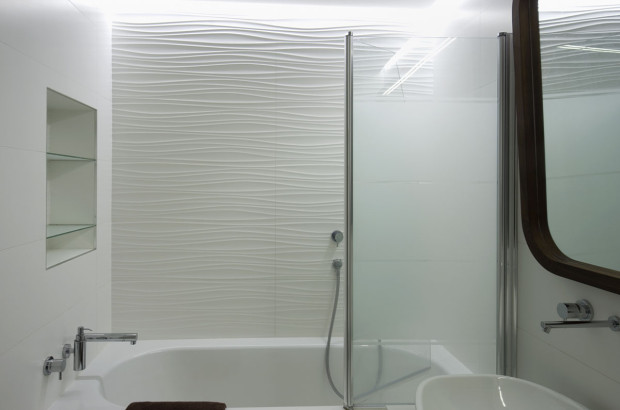 Bílá barva je použita v celém bytě, ani koupelna není výjimkou. Obklad s 3D efektem je zde příjemným ozvláštňujícím momentem − dlaždice s vlnitou strukturou kontrastuje s hladkými povrchy. Vlnitý vzor je zde chápán jako motiv vody.