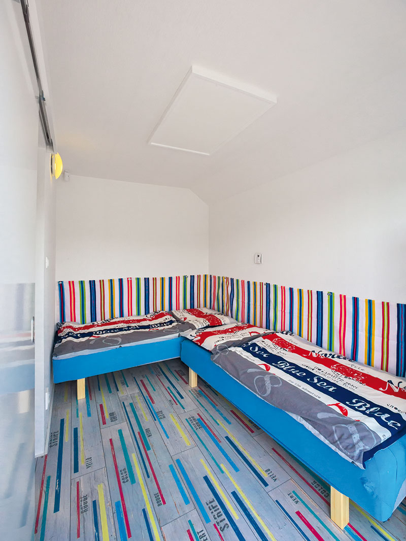 Ložnice jsou v plovoucí chatě dvě – jedna slouží rodičům, druhá dětem. Pro oživení i pohodlné opření je v té dětské za postelemi umístěn pruhovaný látkový sokl v živých barvách. FOTO KATARÍNA BAKO