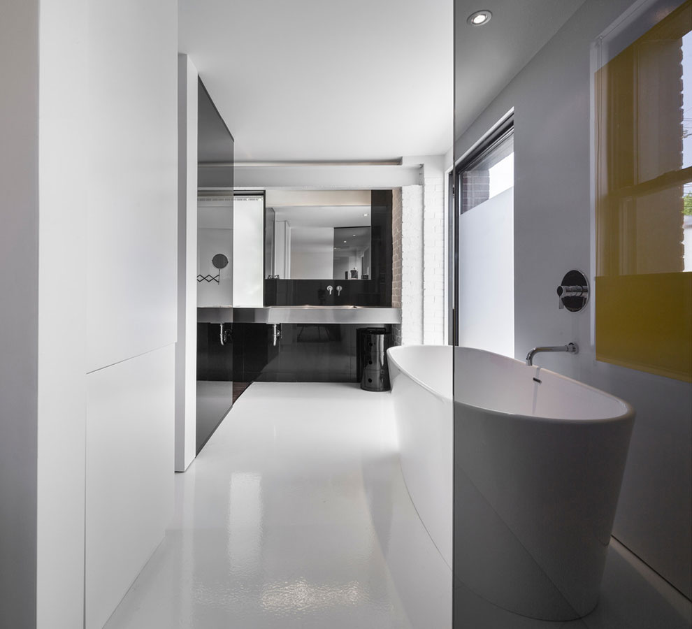 Koupelna je dle moderních trendů pojata jako součást ložnice, tedy řešení en-suite. Cihlový obklad natřený na bílo svou „hrubou strukturou“ narušuje jednolitost hladkých materiálů. Foto: Stéphane Groleau