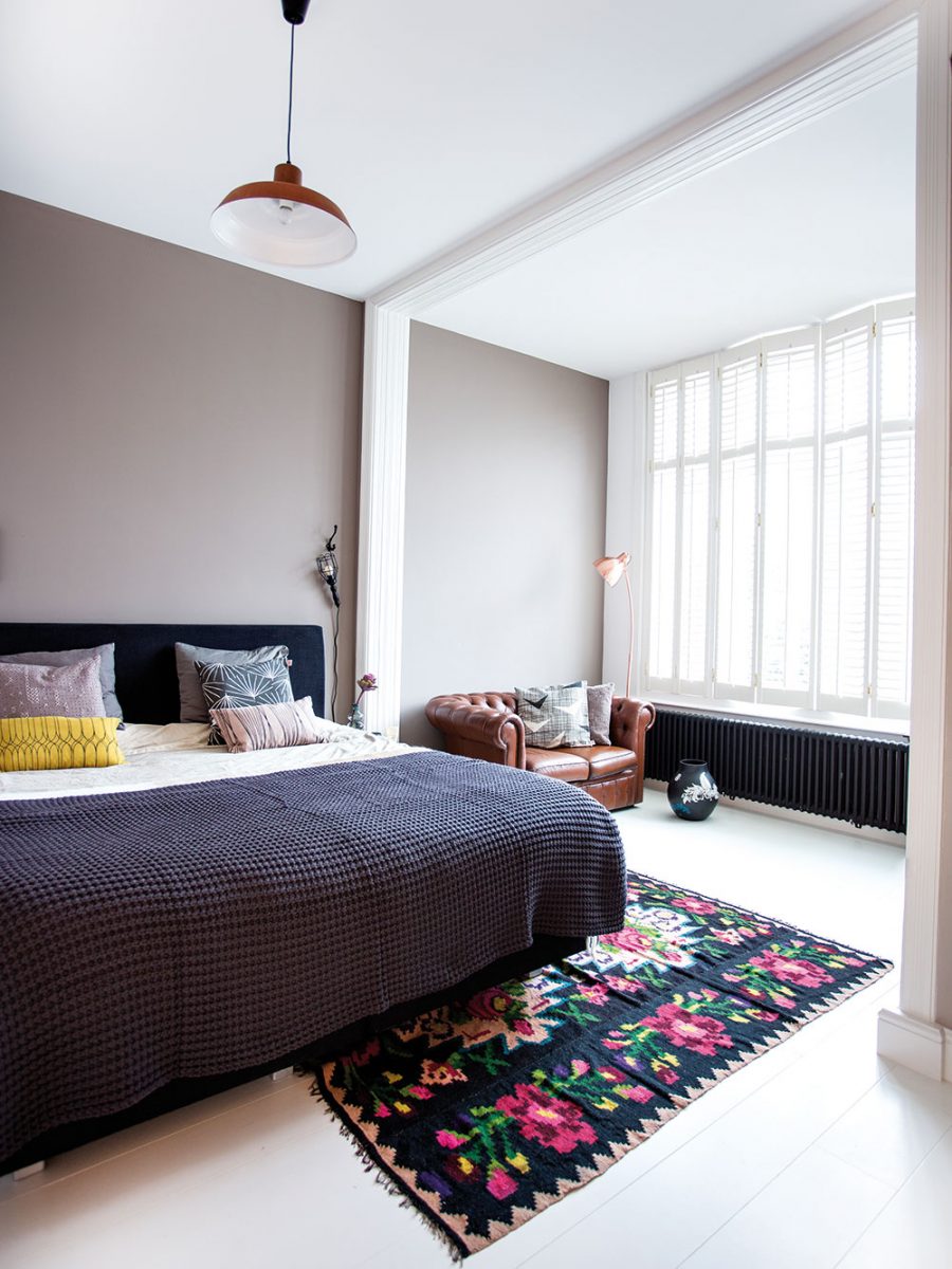Tlumenou šedou zeď v ložnici doplňuje pestrobarevný květovaný koberec a vzorované polštáře. Foto Femque Schook pro Westwing Home and Living