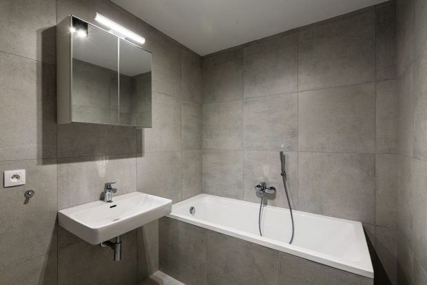 Jednoduché čtvercové obklady šedé barvy pokrývají v koupelně podlahu, stěny i vanu. Nenápadný vzhled místnosti doplňuje celkový charakter bytu. FOTO FRANTIŠEK GÉLA, FABIÁN FRONČEK