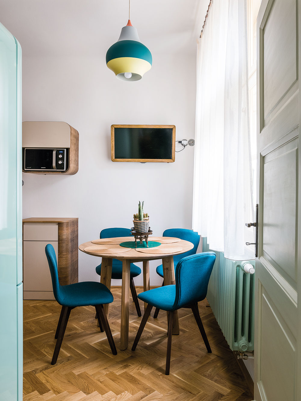 Součástí kuchyně je i sympatický jídelní koutek s modrými pohodlnými židlemi a retro lampou v podobném odstínu. Majitelé však nejraději jedí v prostorném obývacím pokoji. FOTO NORA A JAKUB ČAPRNKOVI