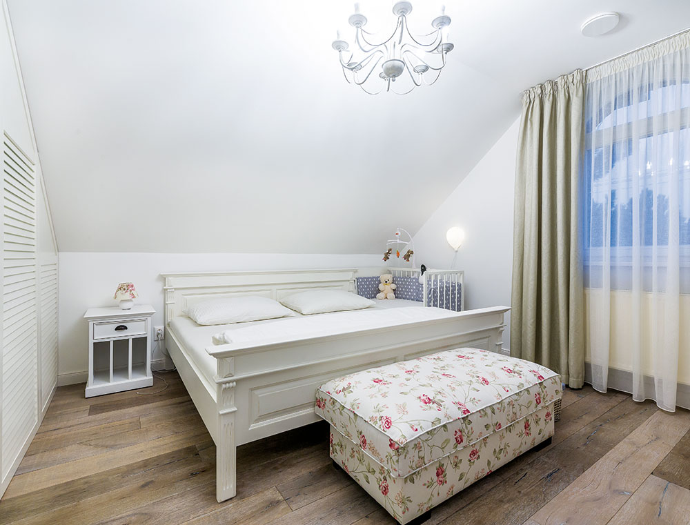 Bílá masivní postel a podnožka s romantickým květinovým potahem tvoří kontrast s dřevěnou podlahou, vše to působí útulně a čistě. Dojem elegantní ložnice na anglický způsob se vydařil. FOTO MIRO POCHYBA