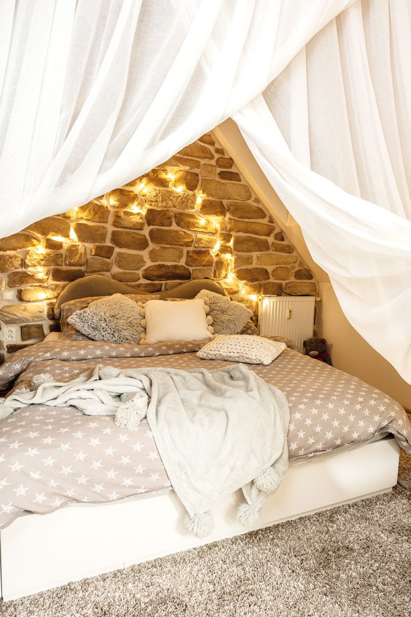 Romanina ložnice je umístěna nad kuchyní a stěna v záhlaví postele je také obložena kameny. Bílé závěsy přispívají k pocitu bezpečného hnízda. FOTO JIŘÍ HURT
