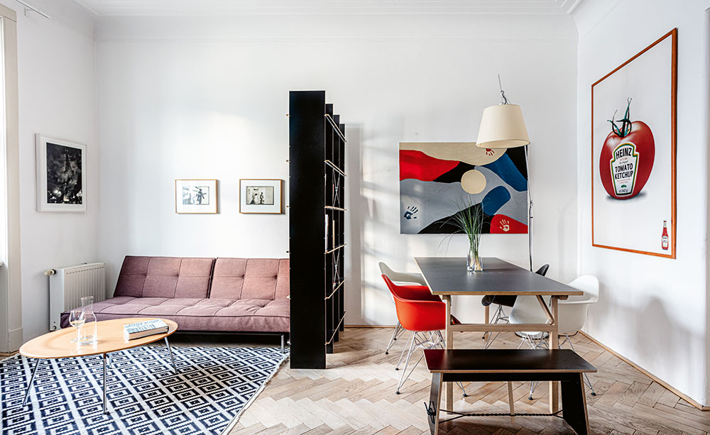 Prostor obývacího pokoje je rozdělený oboustranným otevřeným regálem z produkce značky Nils Hilger Moormann –  stejně jako velký jídelní stůl s nastavitelnými nohami. Střídmé moderní vybavení doplňuje ikonická stojací lampa Tolomeo a židle eamsovky. FOTO JIŘÍ HURT