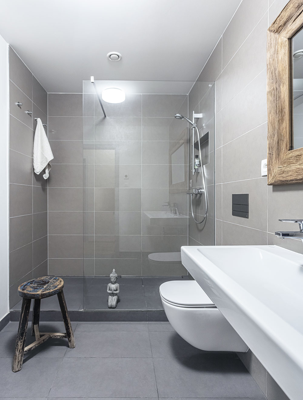 Koupelna je obložená šedým obkladem a se zbytkem bytu ji stylově spojují dřevěné zařizovací prvky, jako je rám zrcadla nad umyvadlem a praktická stolička. FOTO JIŘÍ HURT