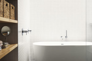 Bílé obložení stěn doplňuje černá mozaiková dlažba a přírodní dřevo na vodorovných plochách koupelny