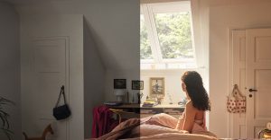 Mladá žena sedí na posteli a dívá se z okna.