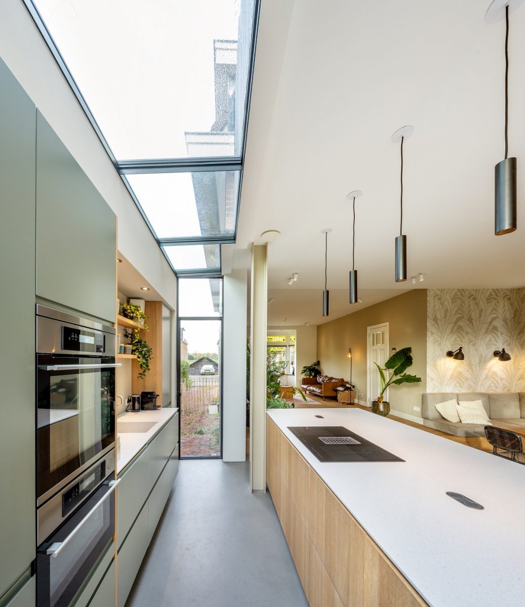 Interiér moderní kuchyně s vysokými stropy a střešními okny, skrz která prosvítá denní světlo. Na levé straně je vestavěná kuchyňská linka v odstínech šedé s vestavěnými spotřebiči. 