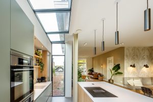 Interiér moderní kuchyně s vysokými stropy a střešními okny, skrz která prosvítá denní světlo. Na levé straně je vestavěná kuchyňská linka v odstínech šedé s vestavěnými spotřebiči.