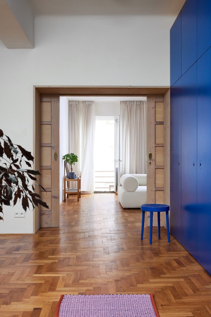 Chodba s dřevěnou podlahou, modrými skříňkami a výhledem do světlého obývacího pokoje.