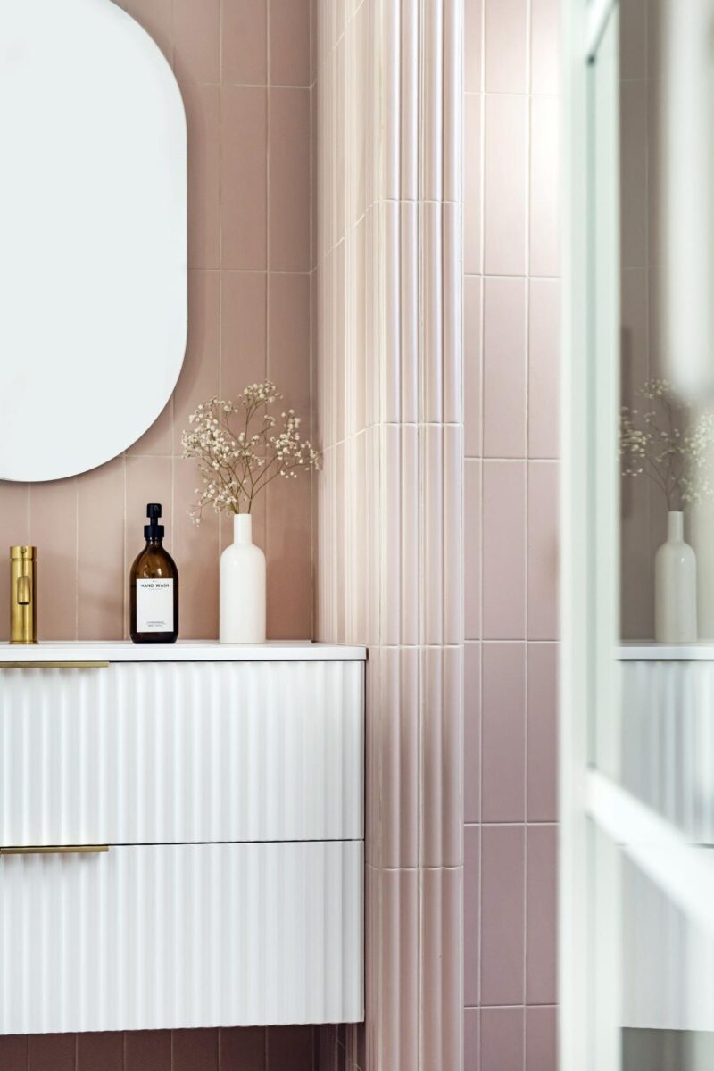 Elegantní koupelna s růžovými dlaždicemi a bílou skříňkou pod oválným zrcadlem.