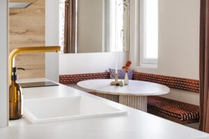 Moderní kuchyň s bílým umyvadlem, zlatou baterií a výhledem na jídelní kout s kulatým stolem.