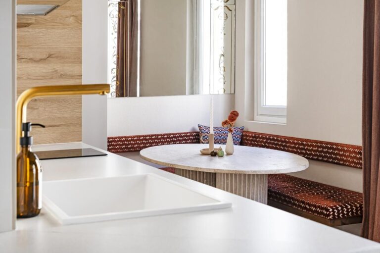 Moderní kuchyň s bílým umyvadlem, zlatou baterií a výhledem na jídelní kout s kulatým stolem.