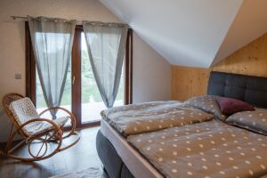 Šedá postel s vysokým čalouněným čelem, houpací křeslo a světlé koberce představují minimalistické, avšak zcela dostačující vybavení pokoje.