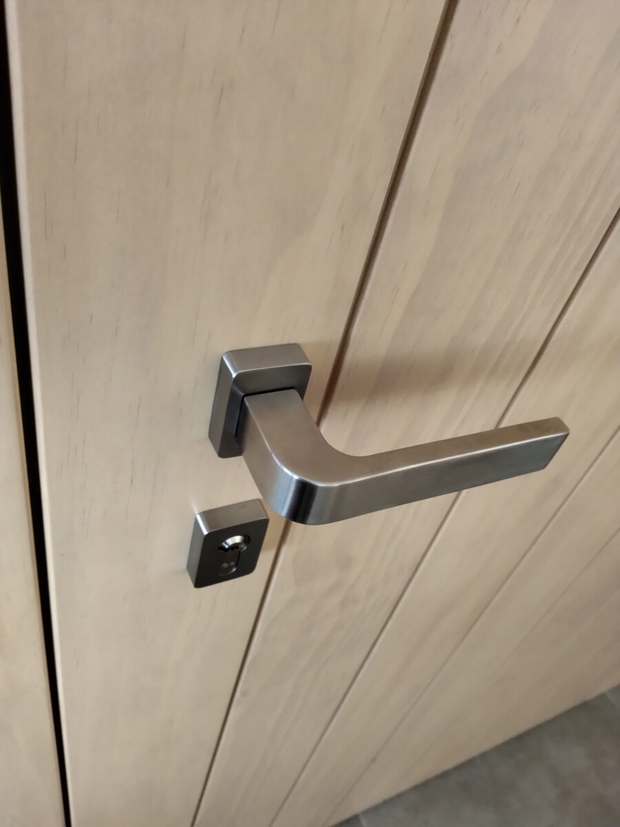 Minimalistická nerezová klika a zámek na světle dřevěných dveřích v moderním interiéru.