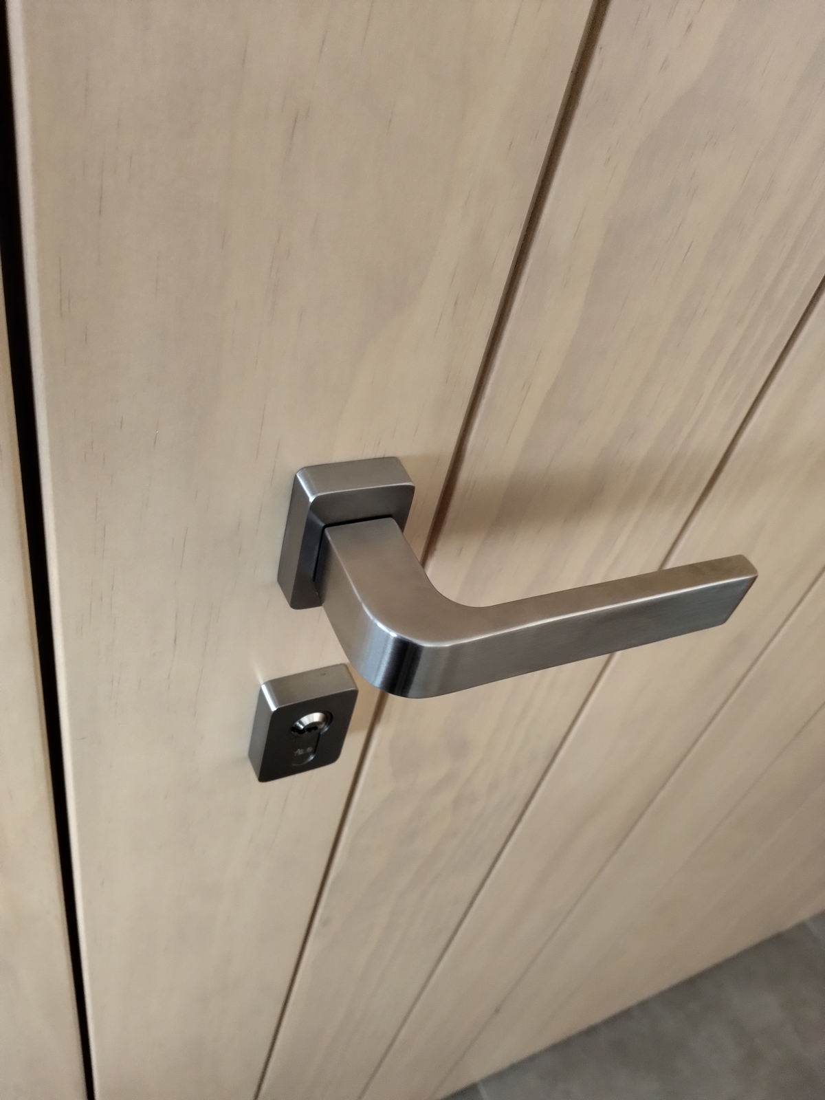 Minimalistická nerezová klika a zámek na světle dřevěných dveřích v moderním interiéru.