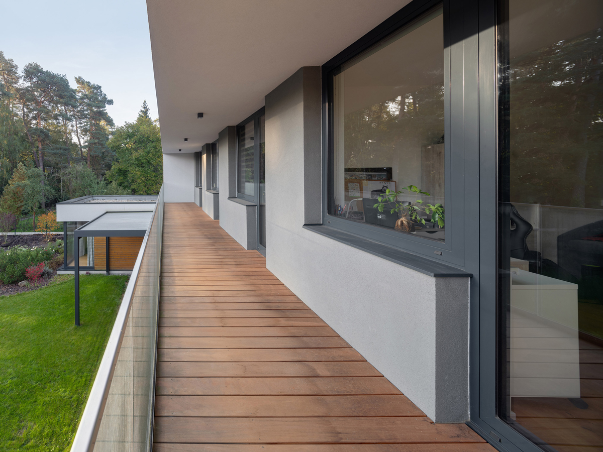 Dlouhý dřevěný balkon s proskleným zábradlím podél patra vily, nabízející klidný výhled do okolní přírody a na moderní přístavbu.
