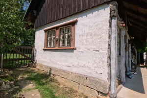 Boční stěna bílé roubenky s typickým malovaným oknem, kamenný základ a čerstvá tráva kolem.