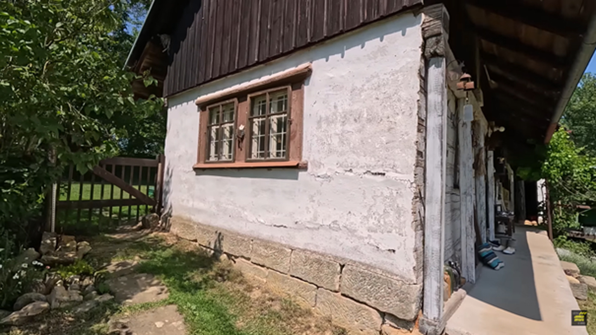 Boční stěna bílé roubenky s typickým malovaným oknem, kamenný základ a čerstvá tráva kolem.