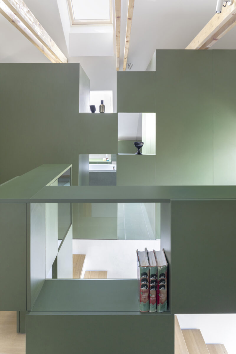 Minimalistická zelená knihovna s geometrickým členěním a průhledy, která rozděluje podkrovní prostor, v pozadí dřevěné schodiště.