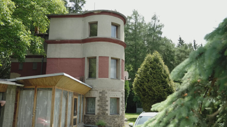 Pohled na zaoblený roh dvoupodlažní vily s červenobílou fasádou a přístavkem, obklopenou zelení.