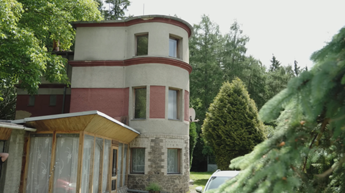 Pohled na zaoblený roh dvoupodlažní vily s červenobílou fasádou a přístavkem, obklopenou zelení.