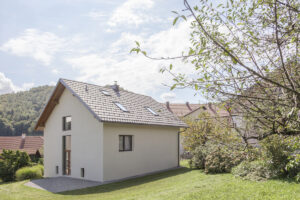 Exteriér rodinného domu s bílou fasádou, sedlovou střechou a střešními okny, zasazeného v zahradě na úpatí lesa.