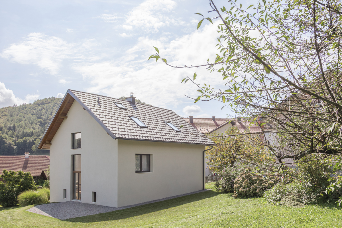 Exteriér rodinného domu s bílou fasádou, sedlovou střechou a střešními okny, zasazeného v zahradě na úpatí lesa.