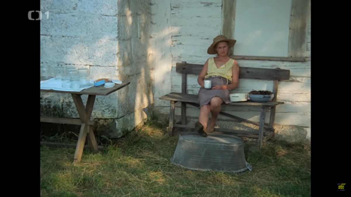 Žena v klobouku sedí na lavici před chalupou, popíjí kávu a má nohy položené na kovovém lavoru, vedle ní je stůl s nádobím a mísa se švestkami.