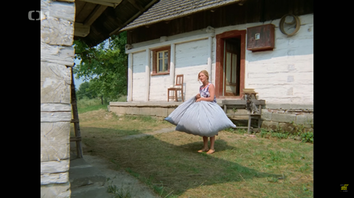Filmová scéna: Žena s velkou peřinou před bílou roubenou chalupou Titulek: Slavná scéna z filmu, kdy Marie vynáší peřiny na zápraží chalupy.