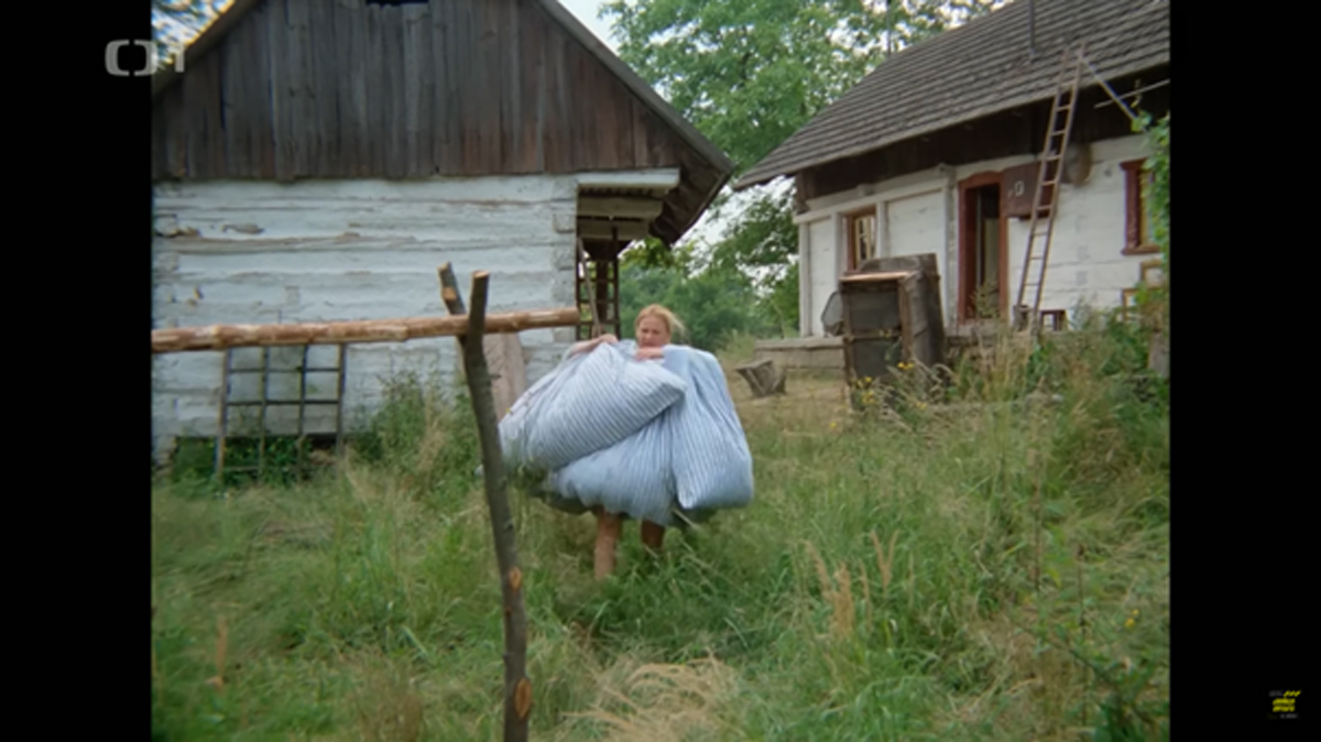 Filmová scéna: Žena nese několik peřin přes zarostlý dvůr mezi chalupami