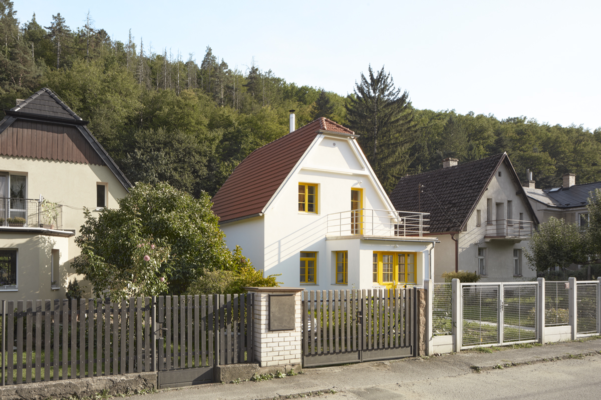 Fasáda bílé vily s výraznými žlutými rámy oken a střechou z červených tašek, zasazená do klidné ulice u lesa.
