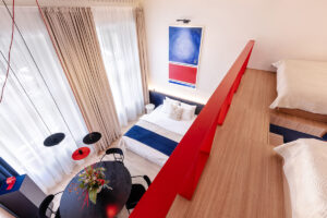 Pohled shora na dvoupodlažní apartmán s manželskou postelí, kulatým jídelním stolem, designovým osvětlením a výrazným obrazem na stěně.