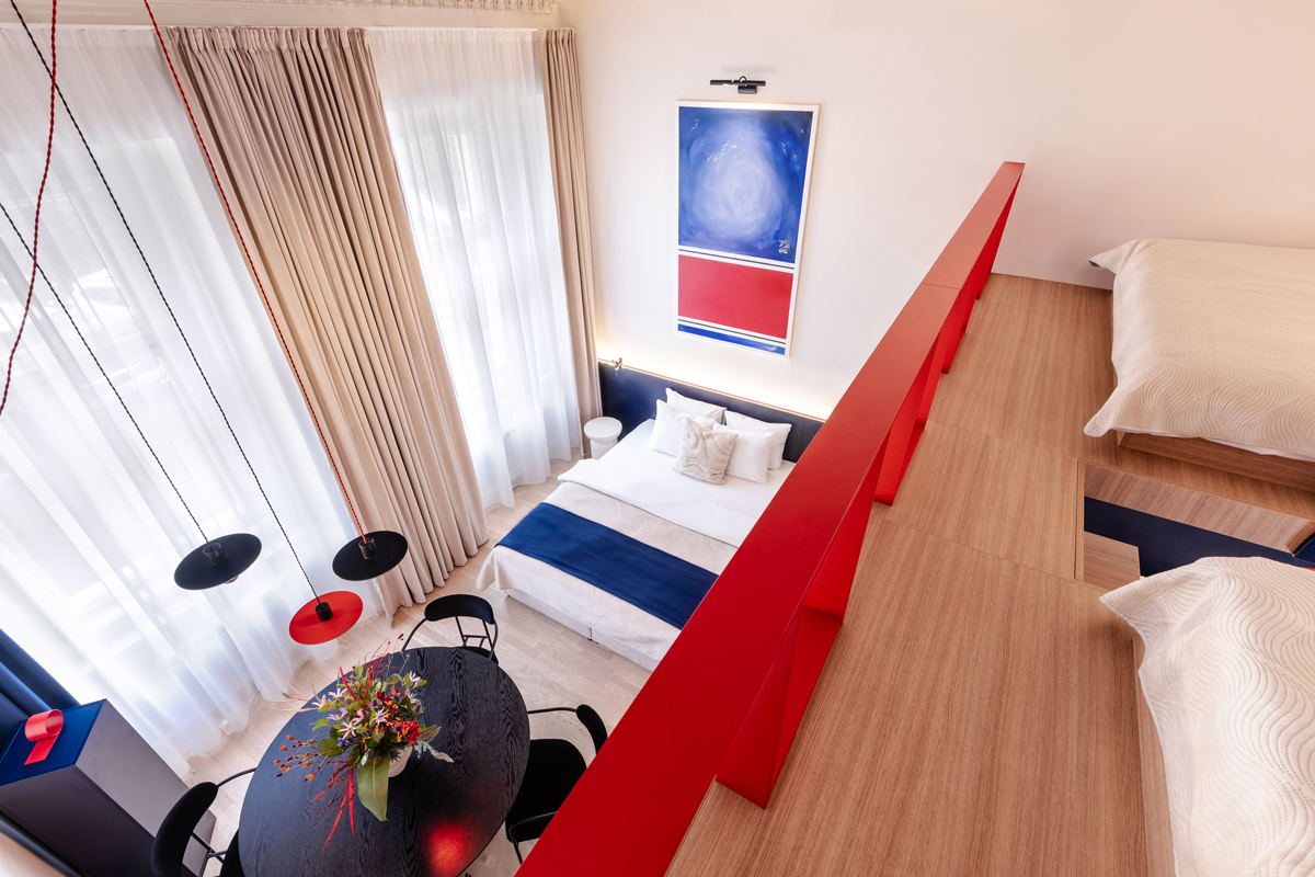 Pohled shora na dvoupodlažní apartmán s manželskou postelí, kulatým jídelním stolem, designovým osvětlením a výrazným obrazem na stěně.