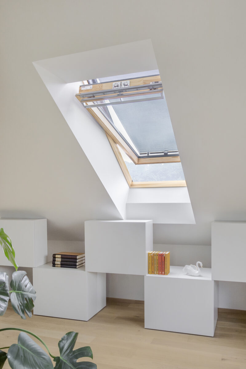 Otevřené střešní okno s roletou v moderním bílém interiéru, kde pod ním stojí minimalistický nábytek s knihami a porcelánovou labutí.
