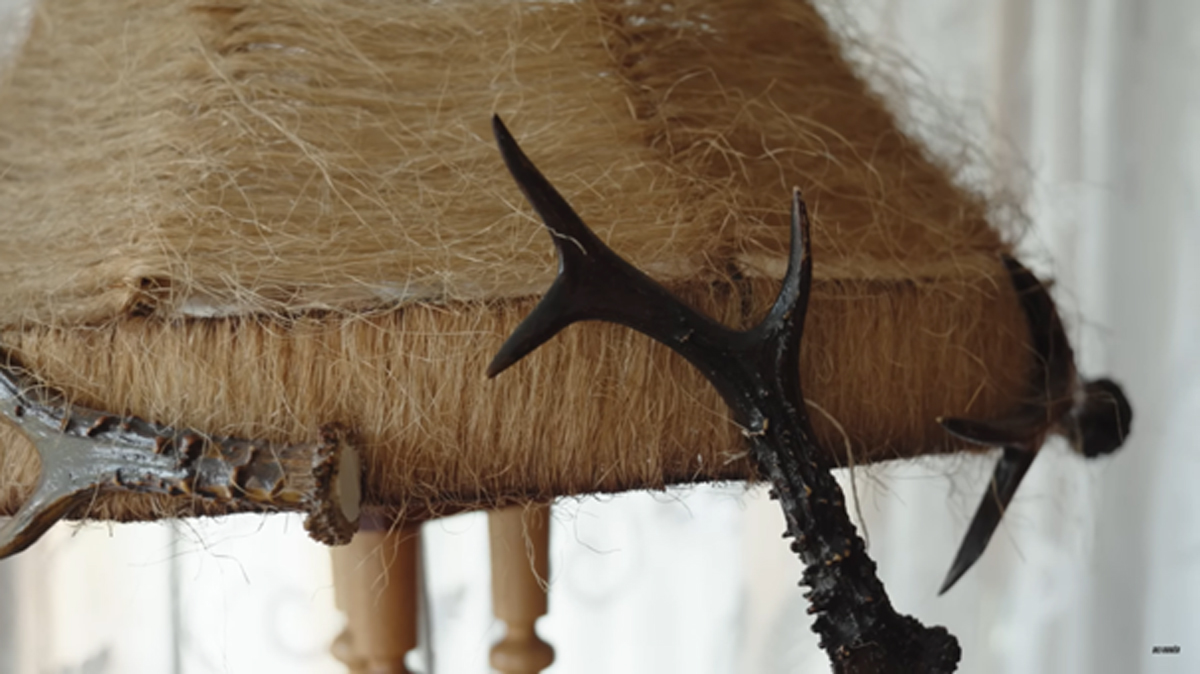 Detail lampy zdobené parožím a přírodním vláknem, působící jako lovecký artefakt.