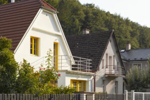 Moderní vila s bílou fasádou, žlutými okenními rámy a červenou střechou za tmavým plotem, obklopená zelení a sousedními domy.