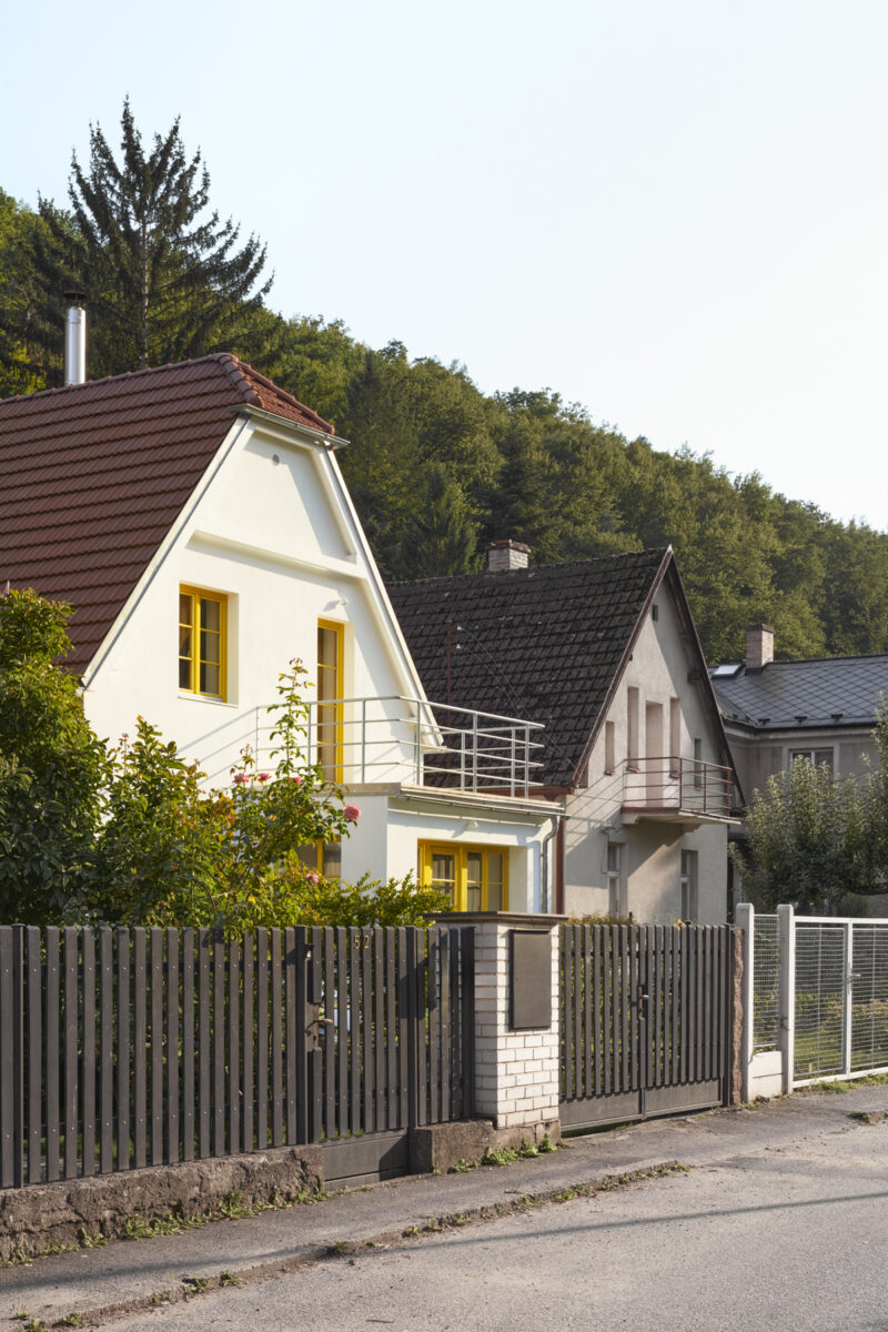 Moderní vila s bílou fasádou, žlutými okenními rámy a červenou střechou za tmavým plotem, obklopená zelení a sousedními domy.