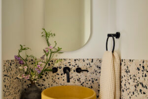 Kompaktní koupelna s terrazzo obkladem, žlutým umyvadlem a minimalistickým zrcadlem.