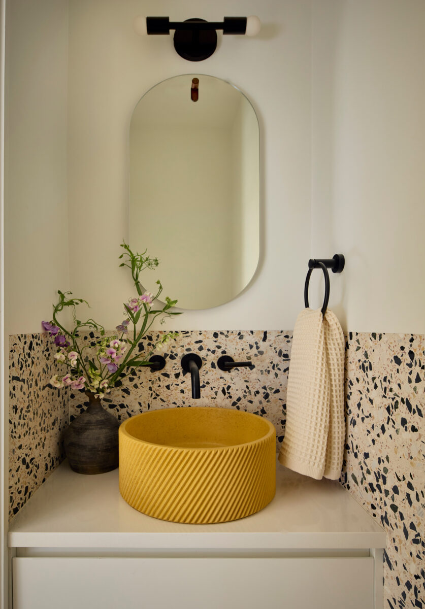 Kompaktní koupelna s terrazzo obkladem, žlutým umyvadlem a minimalistickým zrcadlem.