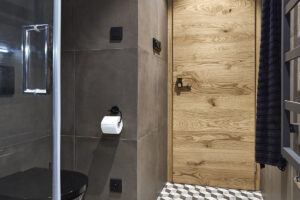 Detailní pohled na vstup do koupelny, dubové dveře, tmavé stěny a černobílá geometrická dlažba tvořící výrazný kontrast.