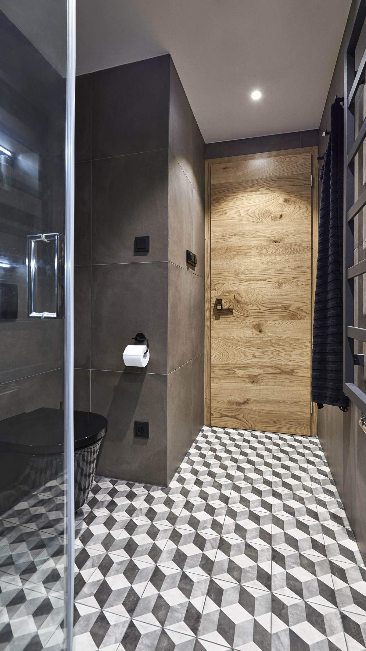 Detailní pohled na vstup do koupelny, dubové dveře, tmavé stěny a černobílá geometrická dlažba tvořící výrazný kontrast.