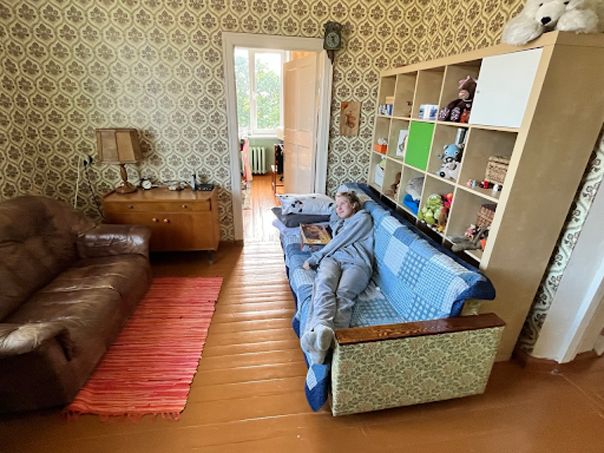 Obývací pokoj v retro stylu s květovanou tapetou, pohovkou a dívkou ležící na gauči s dekou, vedle regálu s hračkami a dekoracemi.