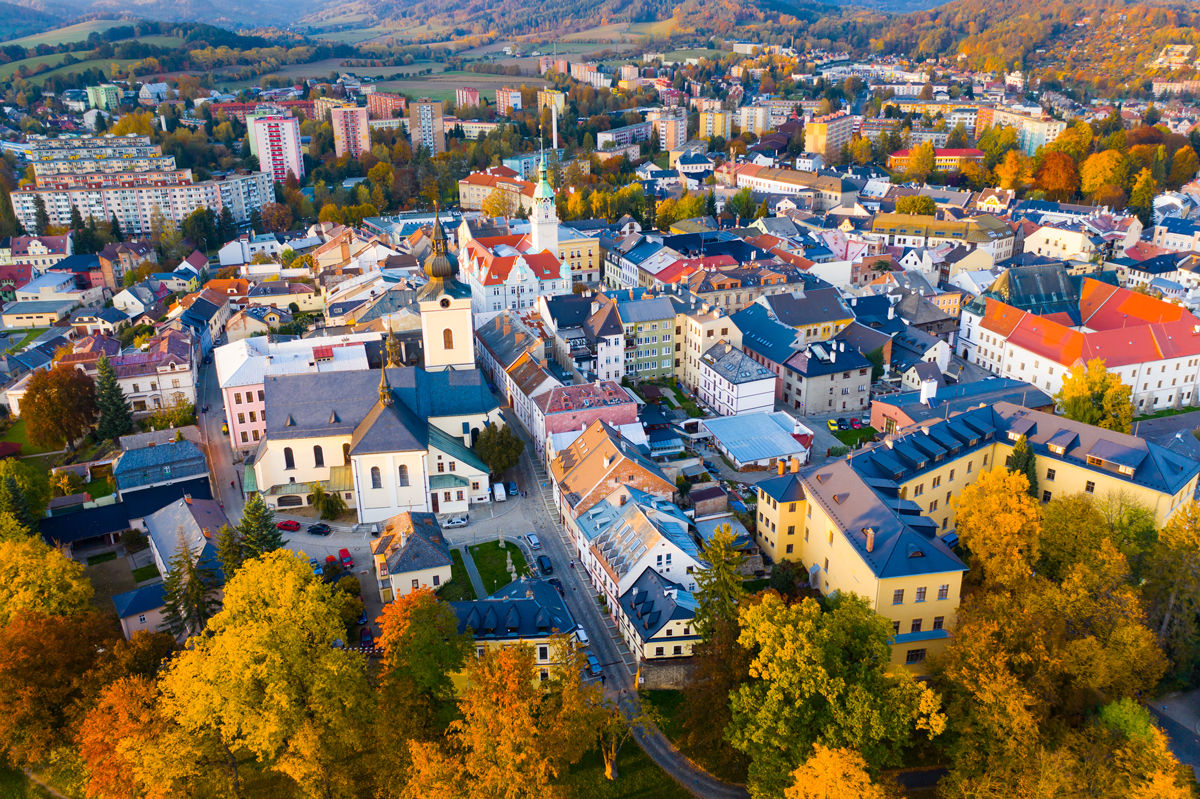 Letecký pohled na centrum českého města s kostelem, barevnými střechami domů a podzimní krajinou v okolí.