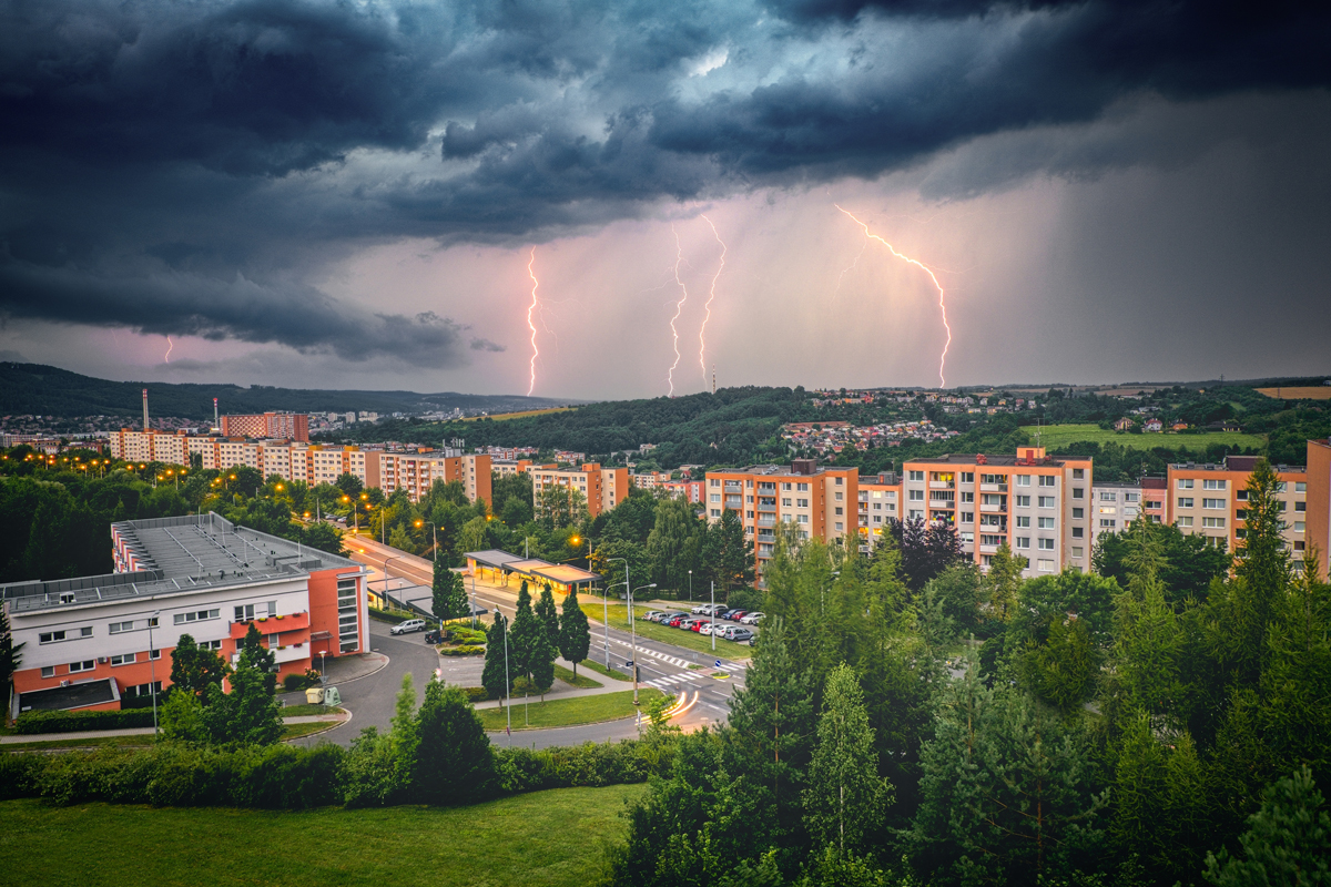 Pohled na sídliště během bouřky, tmavé mraky a několik blesků nad krajinou, v popředí panelové domy a zeleň.